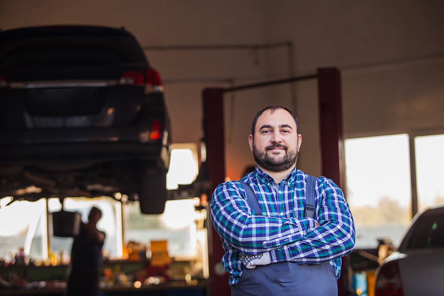 Business Insurance - Portrait of a Small Business Owner Standing in His Garage Repair Shop with His Arms Folded with Views of a Mechanic Working on a Car Repair in the Background