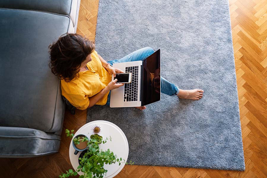 Resources - View From Above of a Young Woman Sitting on a Rug in the Living Room Next to the Sofa While Using a Laptop and Cellphone